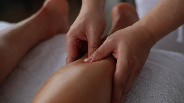 Tracking shot of unrecognizable male masseur doing sports massage to calf muscles of sportsman lying on massage table. Professional therapist with strong hands performing foot and lower leg massage.