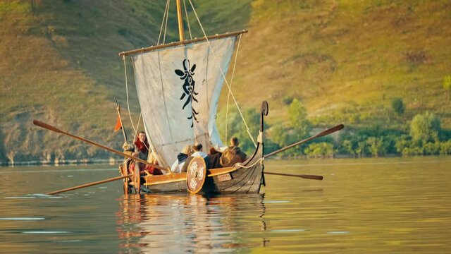Vikings Sail on an Old Ship with a Lowered Sail on a Calm River Against the Backdrop of Rocky Mountains. Concept on the theme of the Vikings and the early Middle Ages.