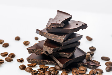 A stack of dark cracked chocolate and black coffee beans on a white background. Pieces of bitter chocolate. Food photography. Close-up.