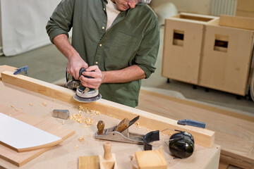 Cropped male carpenter carefully smoothing wooden material with electrical sander, concentrated on...