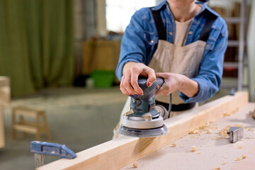 Young cropped female carpenter working with wood using electric sander in workshop. Joinery work on...