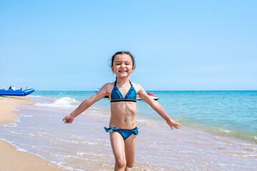 Happy little girl in a bathing suit running with open arms on the golden sand, looking at the camera, spending free time on the beach by the sea. Full length, front view. Childhood concept. Summer vac