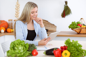 An attractive young blond woman chooses the recipe for a delicious meal while sitting at the table in the kitchen. Tablet pc is the best cookbook