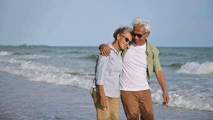 Asian senior couple tourist wear sunglasses embrace talk and walking with point in sea water on beach in summer holiday. Happy family woman, man gray hair elderly older retire relax together in love. 