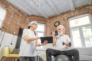 Fototapeta premium Aged woman holding digital tablet while standing on knees on yoga mat. Grey haired man sitting on fitball and training with dumbbells. Online tutorial for domestic workout.