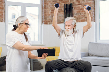 Elderly man training with dumbbells on fitball and watching online tutorial on digital tablet that holding her charming wife in hands. Couple using modern gadgets for workout at home.
