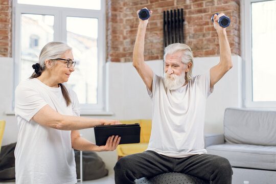 Elderly man training with dumbbells on fitball and watching online tutorial on digital tablet that holding her charming wife in hands. Couple using modern gadgets for workout at home.