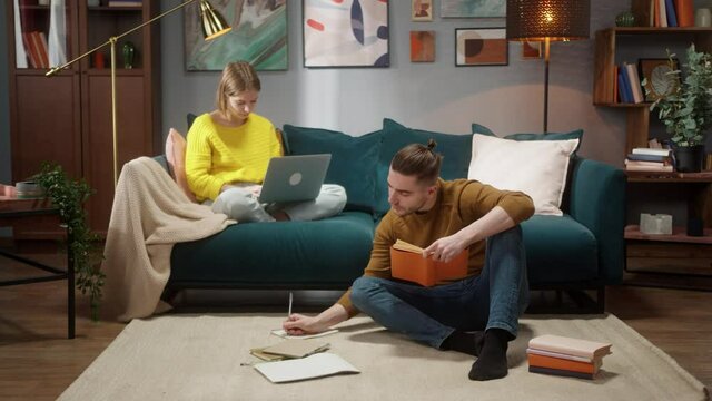 Loving students couple indoors at home preparing for exams, woman using laptop computer. Young family, man writing notes, read homework, remote learning during coronavirus lockdown.