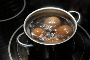 Close-up view of four brown chicken eggs boiling in hot water in small stainless saucepan on black kitchen stove (induction cooktop). Selective focus. Easter preparation theme.