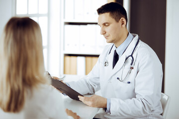 Friendly male doctor and patient woman discussing current health examination while sitting in sunny clinic. Perfect medical service in hospital