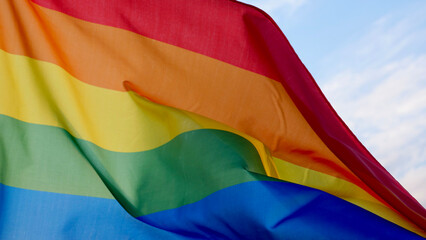 Female hand holding a LGBT gender identity flag on a background of blue cloudy sky on a sunny windy summer day during the celebrating a gay parade, Bisexuality or Coming Out Day