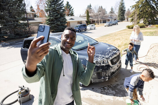 Man taking selfie with family washing car in driveway