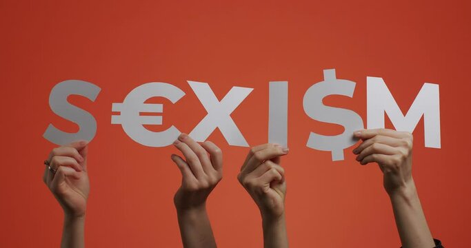 Hands showing word sexism of english language with euro, dollar sign. Word made of carved paper for feminist blog