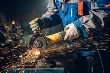 Locksmith in special clothes and goggles works in production. Metal processing with angle grinder. Sparks in metalworking - obrazy, fototapety, plakaty