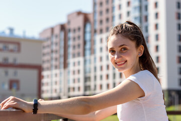 A positive young attractive woman in a white T-shirt and with her hair pulled back in a ponytail poses against the background of new high-rise buildings.Summer day.Urban landscape.Copy space. © Tatyana
