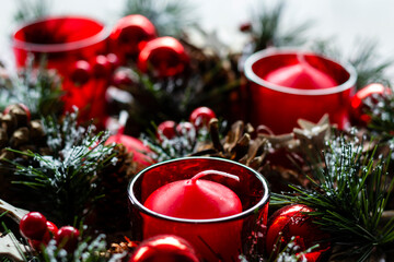 Close view on a decorated Christmas wreath with red candles in red glasses, pine cones and red glass balls. Selective focus on the front candle. Christmas card concept.