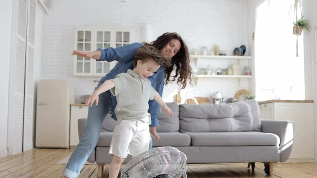 Family bonding at home: playful mother and preschool kid have fun with big indoor fan blow cool wind. Young single parent and child weekend indoor engagement. Cheerful mum enjoy leisure time with son