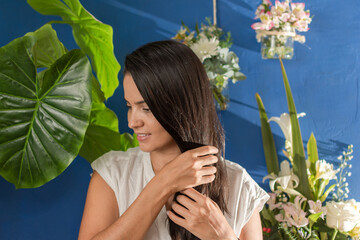 Fashion style beauty portrait of latin woman holding her long shiny black hair and posing between big green tropical leaves and flowers in blue background. Hair care concept