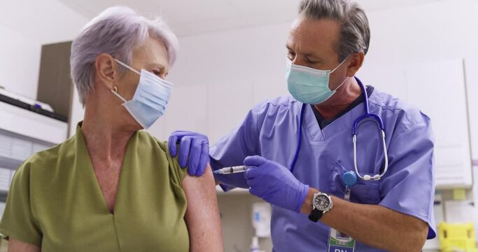 A Mid Shot Of An Australian Male Medical Nurse Administering A Covid-19, SARS-CoV-2 Vaccine Injection With A Needle and Syringe, To An Elderly Woman. Wearing Safety Gloves And Protective Mask