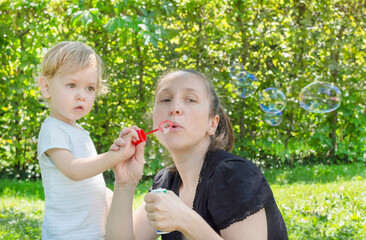 Cute little boy and his mother having fun blowing soap bubbles in the park. Funny outdoor activity for children in a beautiful summer sunny day