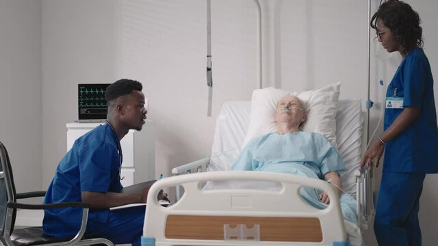 A team of african ethnic group doctors a man and a woman talk to an elderly male patient. A doctor and a nurse talk to a bedridden patient in a hospital ward