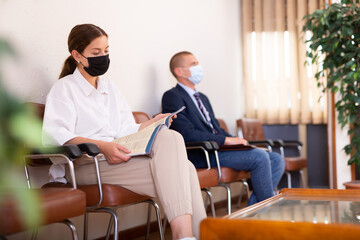 Concentrated girl in a protective mask, sitting in the lobby of a large company waiting for her turn to see a ..specialist during a pandemic, looks at an information magazine