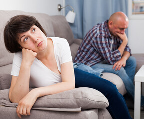 Unhappy mature woman after quarrel, man on background at home interior