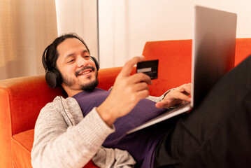 Young man shopping online with credit card at home using computer laying on an orange couch sofa and using headphones
