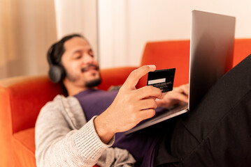 Young man shopping online with credit card at home using computer laying on an orange couch sofa and using headphones