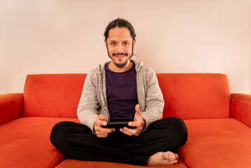Young man playing gaming on the smart phone sitting on an orange sofa couch at home in the living room