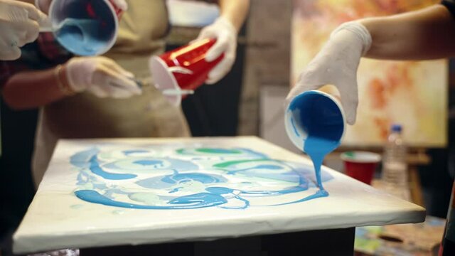 Women artists are pouring acrylic paint on the canvas. Artistic collaboration in workshop.