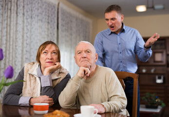 Portrait of frustrated elderly woman and man sitting at home table on background with angry screaming adult son..