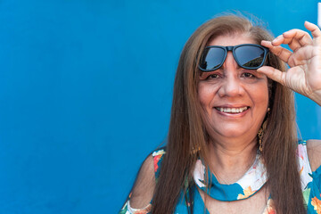 Mature woman holding up her sunglasses. Cheerful woman on blue background.