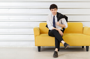 Horizontal portrait shot of young adult Asian handsome man in a white shirt with a suit on the shoulder sitting on the sofa and looking at the camera with hand to chin gesture posing in the studio
