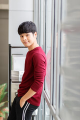 Vertical portrait shot of an attractive smiling young adult Asian man in a casual red shirt standing and leaning against the wall. Young male looking at the camera with hand in pocket posing