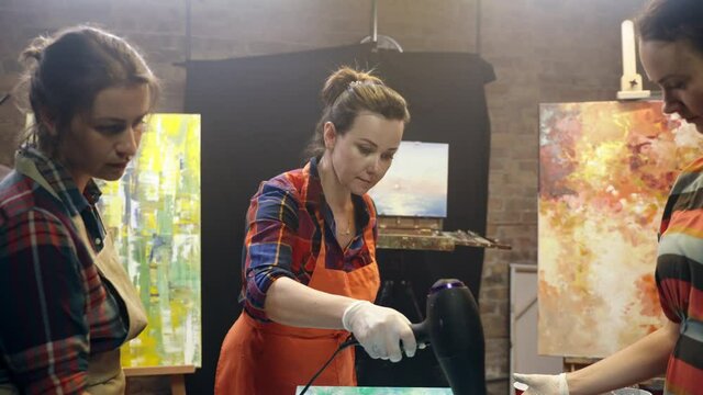 Women artists are drying acrylic paint on the canvas with fan. Artistic collaboration in workshop.
