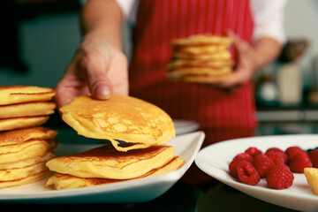 Closeup shot of heap of newly baked yummy fluffy hot golden good smell pancake snack in hand of chef who wears red apron while picking up bread from stack in blurred foreground