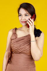 Beautiful lady standing and listening to friend who call her to gossiping about neighbor. Her mobile phone has red color compare with brown dress and yellow background. Photo so colorful.