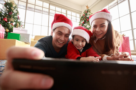 Asian Selfie holding smartphone Taking picture. Happy young family with kids fun celebrating Christmas. Christmas time. My dad, mom, and daughter in Santa hats lie about In front Gift box at home.