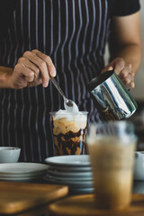 Fototapeta na wymiar Unrecognizable barista using spoon to gentle topping glass of sweet coffee at bar counter with white milk froth from metal jar to enhance taste of softness and mellowness.