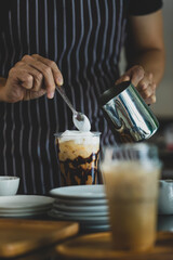 Unrecognizable  barista using spoon to gentle topping glass of sweet coffee at bar counter with white milk froth from metal jar to enhance taste of softness and mellowness.