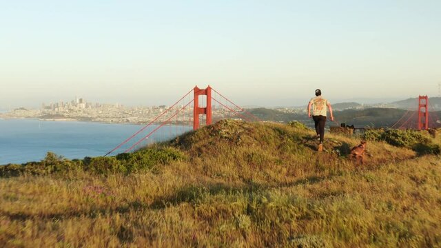 Drone reveal Golden Gate Bridge behind hiker running with dog on hill in sunset