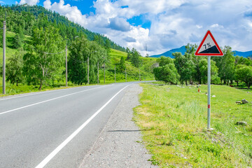 a road sign, a symbol for car drivers, warning about climbing uphill on an asphalt road, on a highway in a picturesque high altitude place in the mountains in cloudy sunny weather