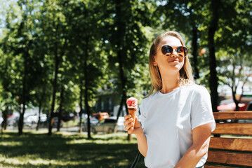 Cheerful, beautiful young woman in glasses holding ice cream and sitting on park bench, summer time. Happy, attractive girl eating waffle cone icecream, enjoying walk outdoors. Copy space. Lifestyle