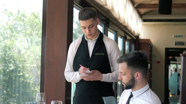 In The Restaurant, The Waiter Takes The Order Of A Stylish Man In An Elite Restaurant. customer service. A waiter who gives recommendations in a restaurant. The client reads the menu