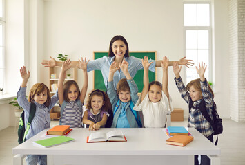 Happy teacher with arms widely spread to hug cheerful children raising hands. Positive portrait in classroom. Educator showing love, adoration and care for pupils. Children and tutor looking at camera - obrazy, fototapety, plakaty