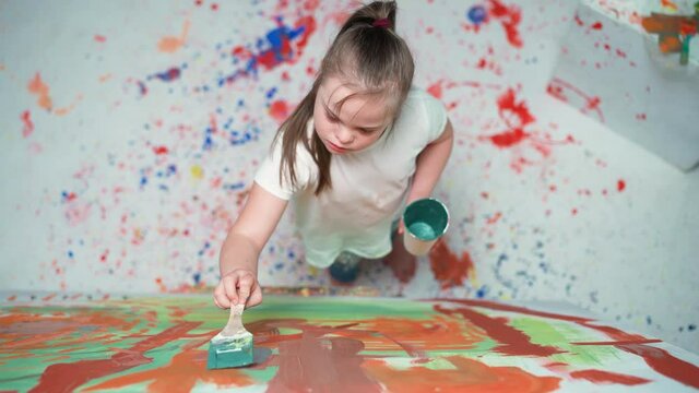 Kid girl with down syndrome draws with a brush on a large canvas in a white room, girl with special needs draws a color abstraction, top view, 4k slow motion.