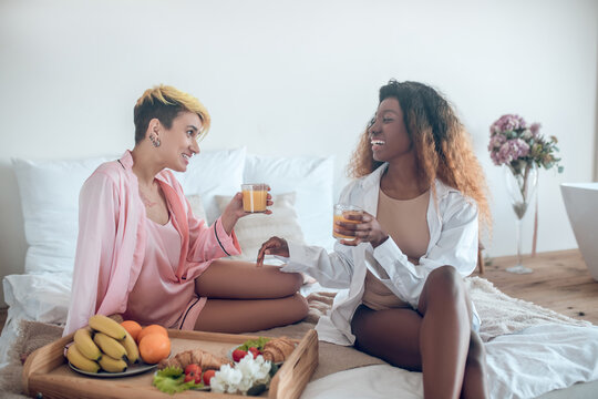 Two laughing women eating on bed in bedroom