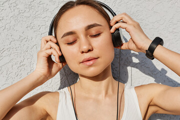 Fototapeta premium Young adult female with dark hair wearing white top, keeping eyes closed, touching headphones with palms, enjoying music after work out, healthy lifestyle.