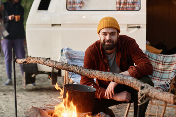 Portrait of young bearded man looking at camera while cooking food on a fire during camping in the forest
